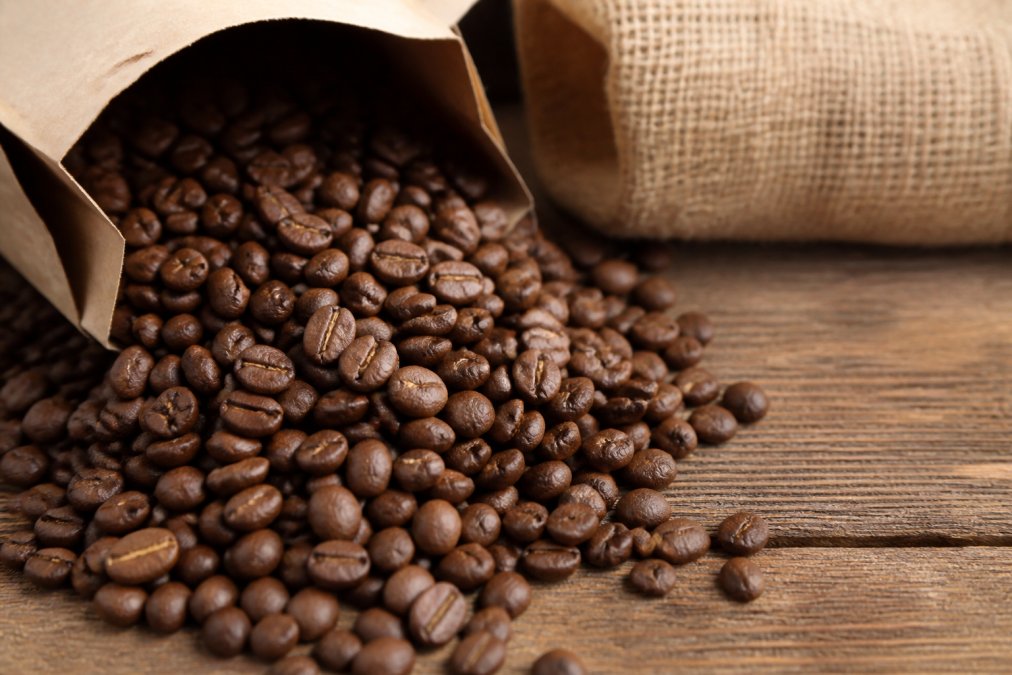 Close-up of whole arabica coffee beans spilling from a kraft paper bag onto a wooden surface, with a roast date stamp visible on the bag label