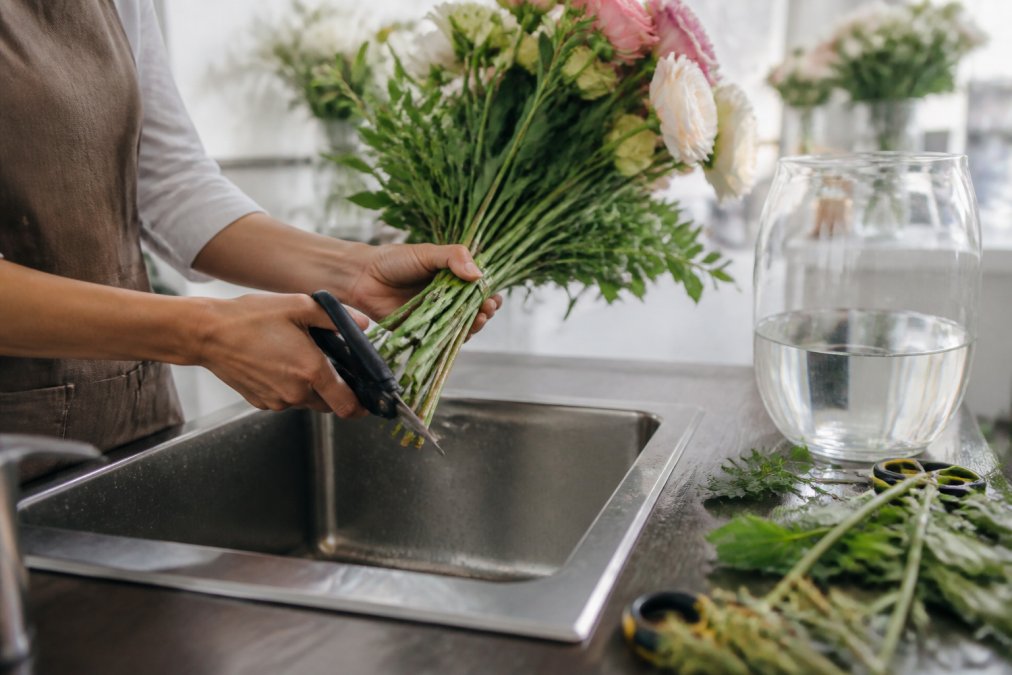 Hands carefully trimming flower stems at a 45-degree angle over a sink with clean water, removing lower leaves from the bouquet before arranging in a clear glass vase