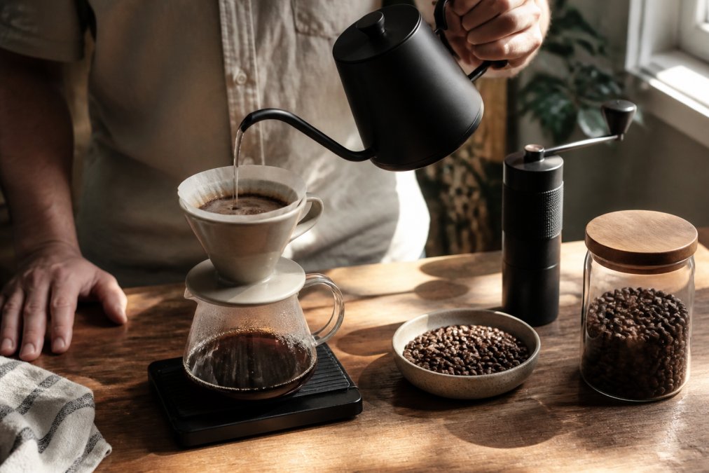 Hands pouring hot water over coffee grounds in a ceramic pour-over dripper, with whole roasted beans and burr grinder visible on wooden surface, morning natural light