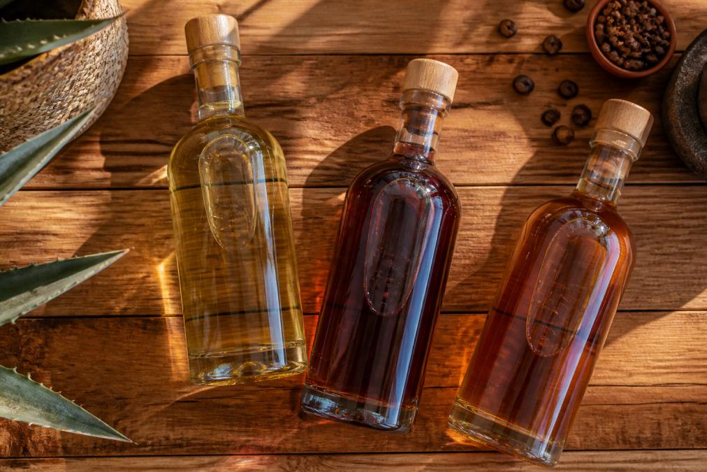 Three premium tequila bottles (Vanilla, Black Cherry, and Coffee varieties) arranged on a wooden surface with natural golden and amber light highlighting the spirits' colours, shot from above with subtle shadow definition