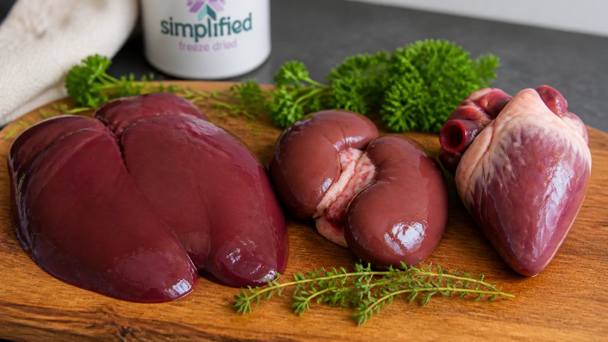 Close-up of raw beef liver, kidney, and heart on a wooden cutting board with fresh herbs, showing the distinct colors and textures of each organ meat