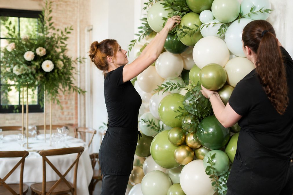 Professional balloon artists installing an organic balloon garland at an event venue, carefully positioning latex and foliage elements with precision while wearing black aprons in a bright indoor setting
