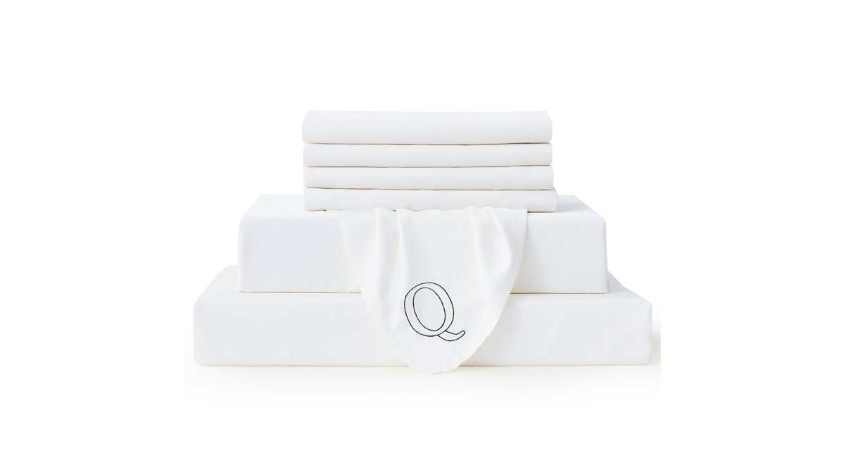 A neatly made queen-size bed in a bright, airy bedroom, crisp white bamboo sheet set with a soft silky sheen, the corner turned back to reveal the embroidered Q indicator