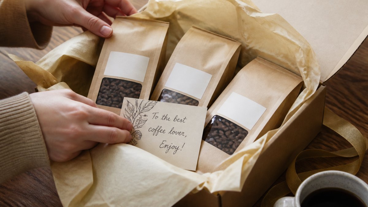 Hands carefully arranging freshly roasted organic coffee sample packs in a premium gift box with cream tissue paper, displaying multiple varieties with personalized handwritten gift card