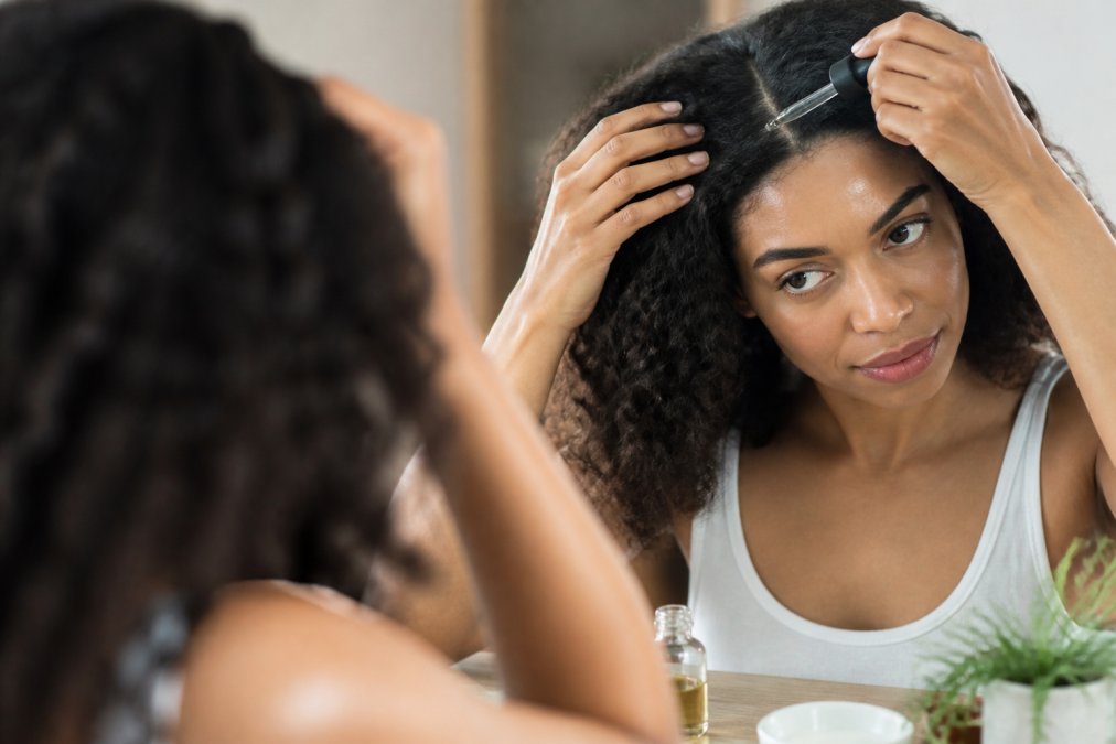 Person with curly hair gently massaging organic hair care oil into their scalp in a bathroom mirror reflection