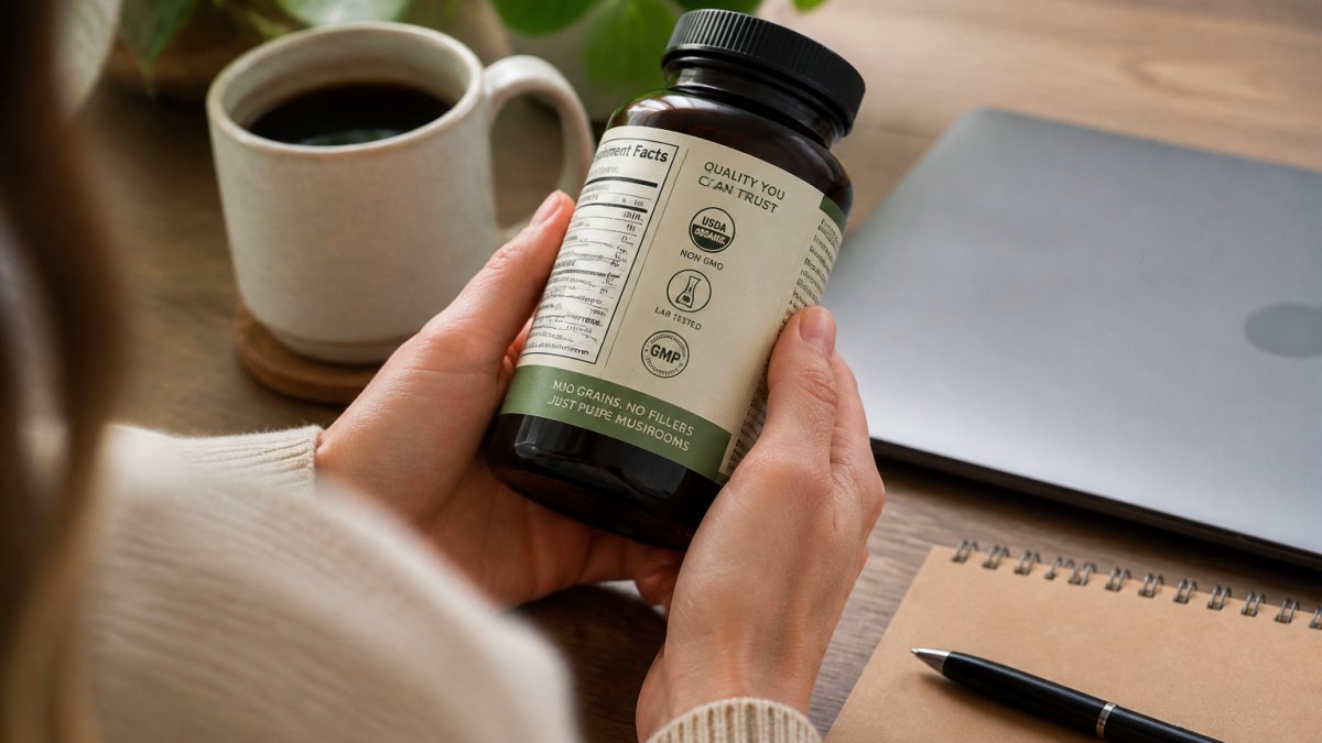 Close-up of hands holding a mushroom supplement bottle next to a laptop and coffee mug on a wooden desk, with a certification label visible on the bottle
