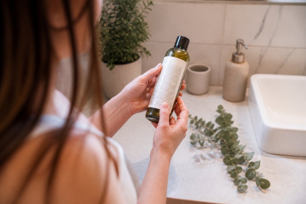 Woman reading the ingredient label on a bottle of organic shampoo in a bright bathroom with eucalyptus.
