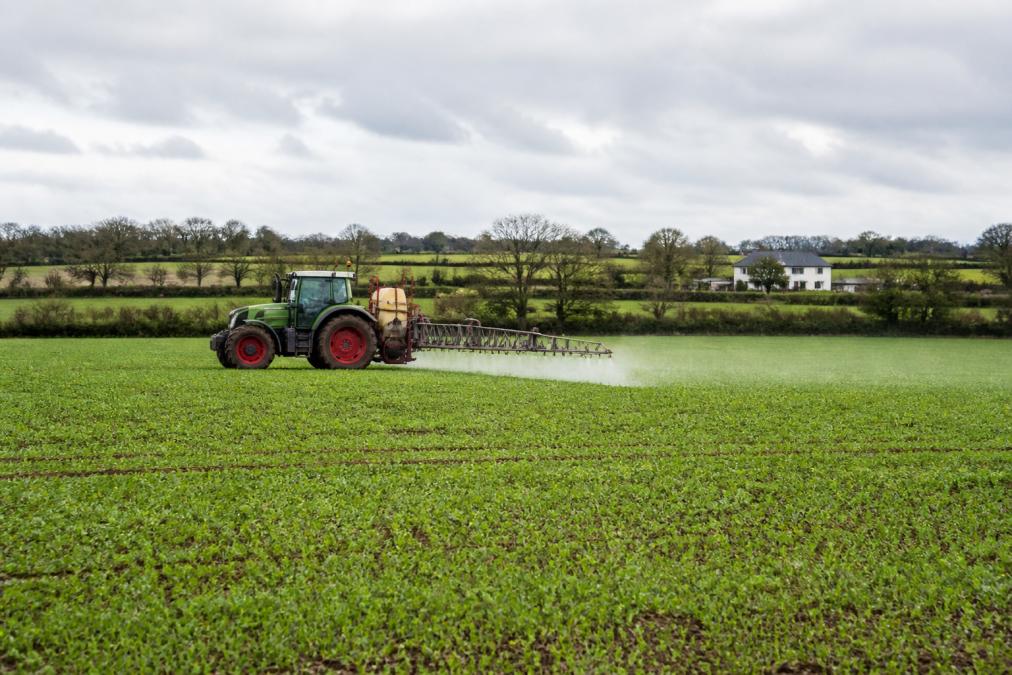 A tractor applying liquid foliar treatment across a large arable field on an overcast day, with a farmhouse and hedgerows visible in the distance