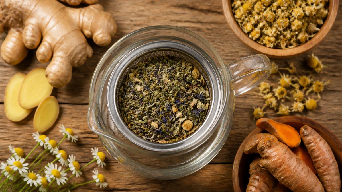 Loose herbal tea leaves in a glass infuser with fresh ginger, chamomile flowers, and turmeric on a wooden table