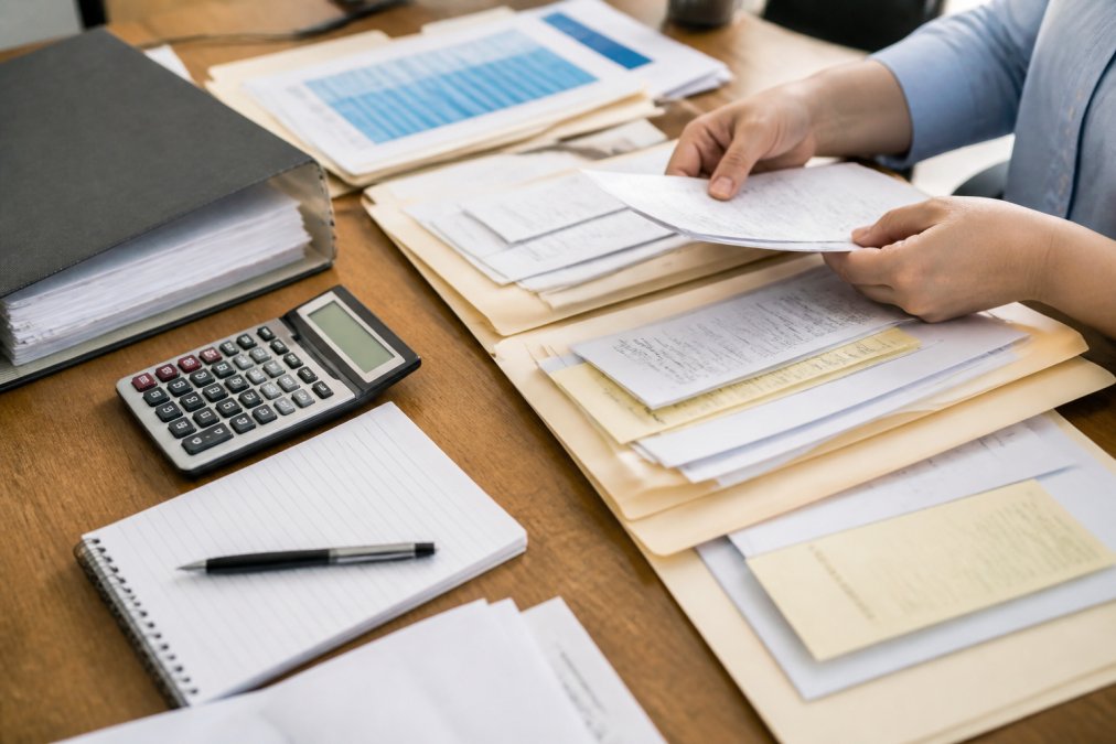 Hands organizing receipts, invoices, and financial documents into labeled folders on a desk with a calculator, notepad, and business ledger visible