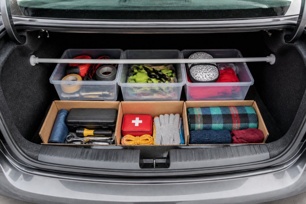 Overhead view of an organized car trunk using clear plastic dollar store bins, tension rods securing items, and labeled cardboard dividers creating compartments for tools, emergency supplies, and seasonal items