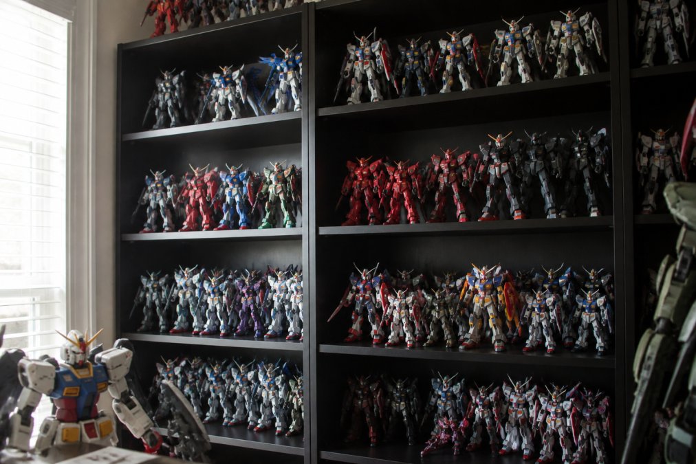 Collector's shelf displaying multiple Gundam model kits arranged by series with varying heights and clear visual separation between different themes, natural window lighting highlighting the details