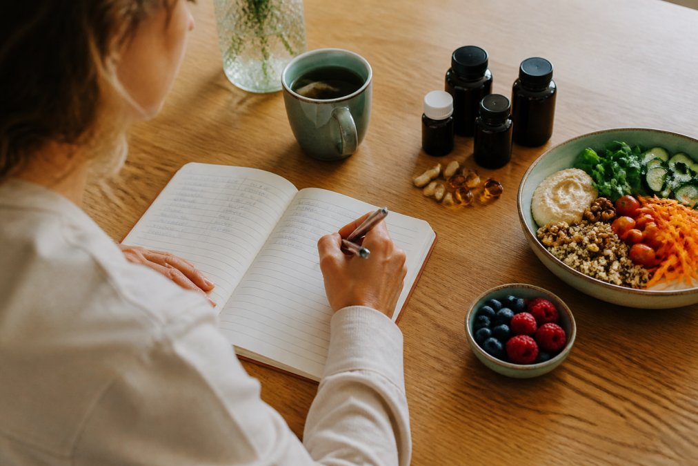 Persoon houdt een notitieboek bij over gezondheid naast orthomoleculaire supplementen, gezonde voeding en een kopje thee.