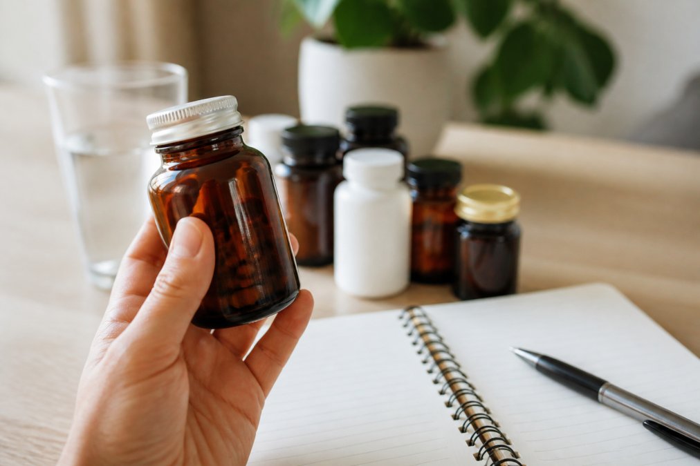 Hand die een pot met supplementen vasthoudt bij een laptop met een medicijnlijst voor orthomoleculaire therapie advies.