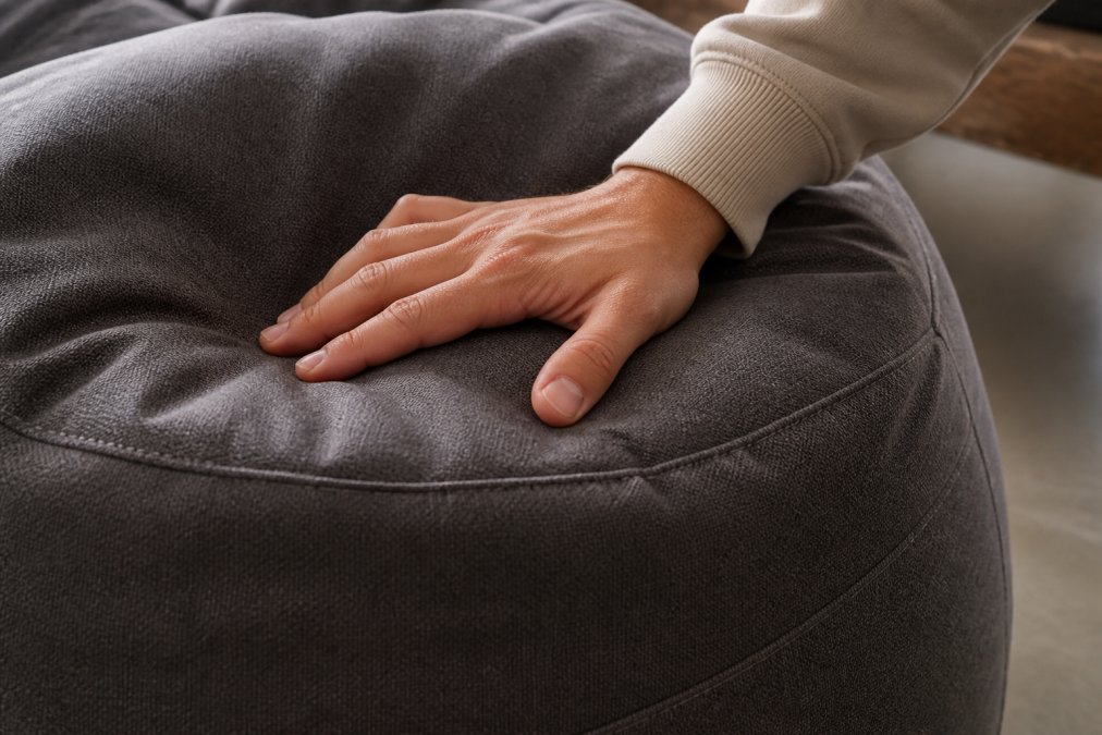 Gros plan sur les détails de construction d'un pouf: coutures serrées, rembourrage épais, base solide, et main touchant le tissu pour montrer la qualité et la durabilité