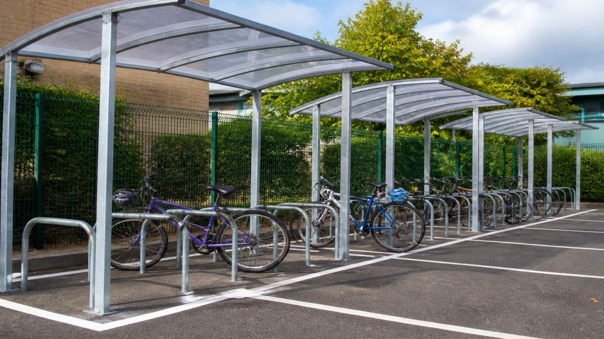 A row of galvanised steel cycle shelters installed in a school car park, bicycles parked under open-sided roofs, bright daylight, tarmac surface with white bay markings