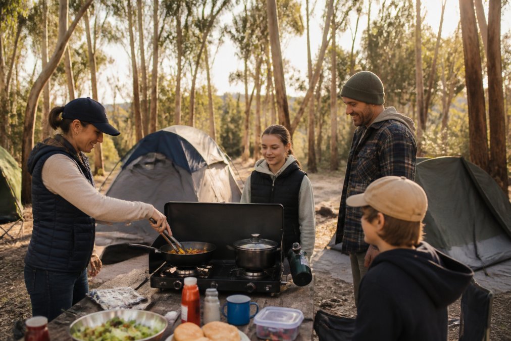A family setting up a portable two-burner camping stove on a picnic table at a campsite, with tents and gum trees in the background under late afternoon light