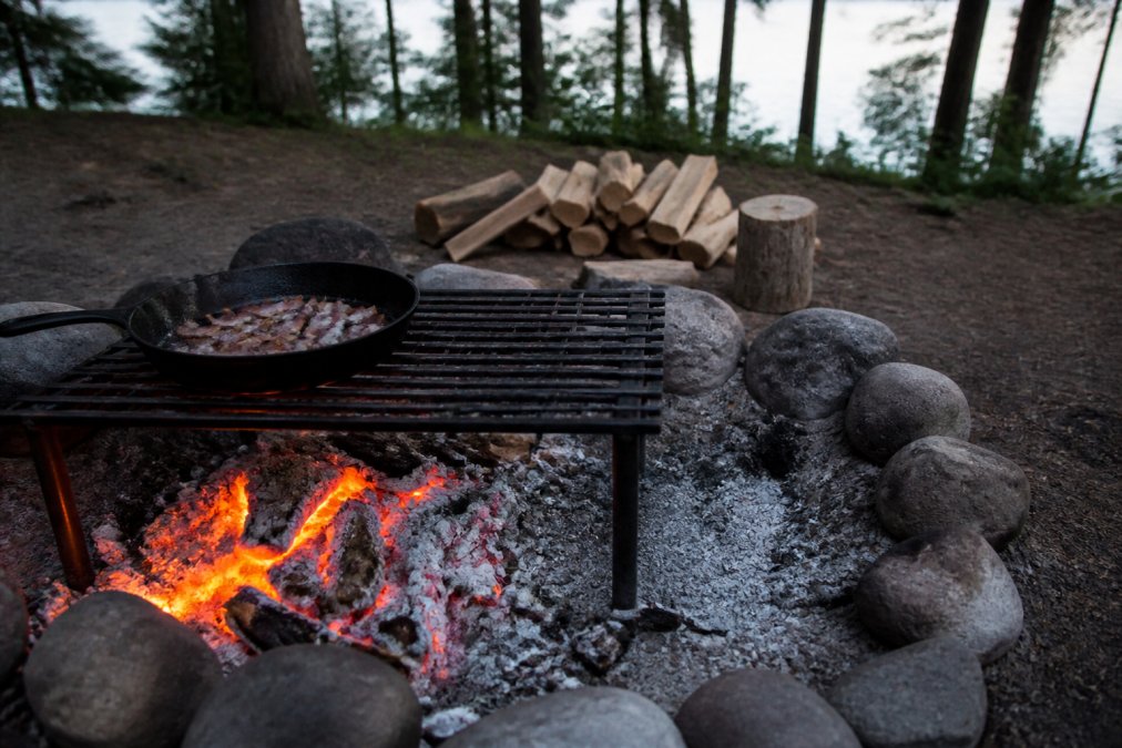 A cast iron skillet with bacon sits on a grate over glowing orange coals, with a clear cooler zone at the edge of the fire ring for temperature control, evening light in a forest clearing