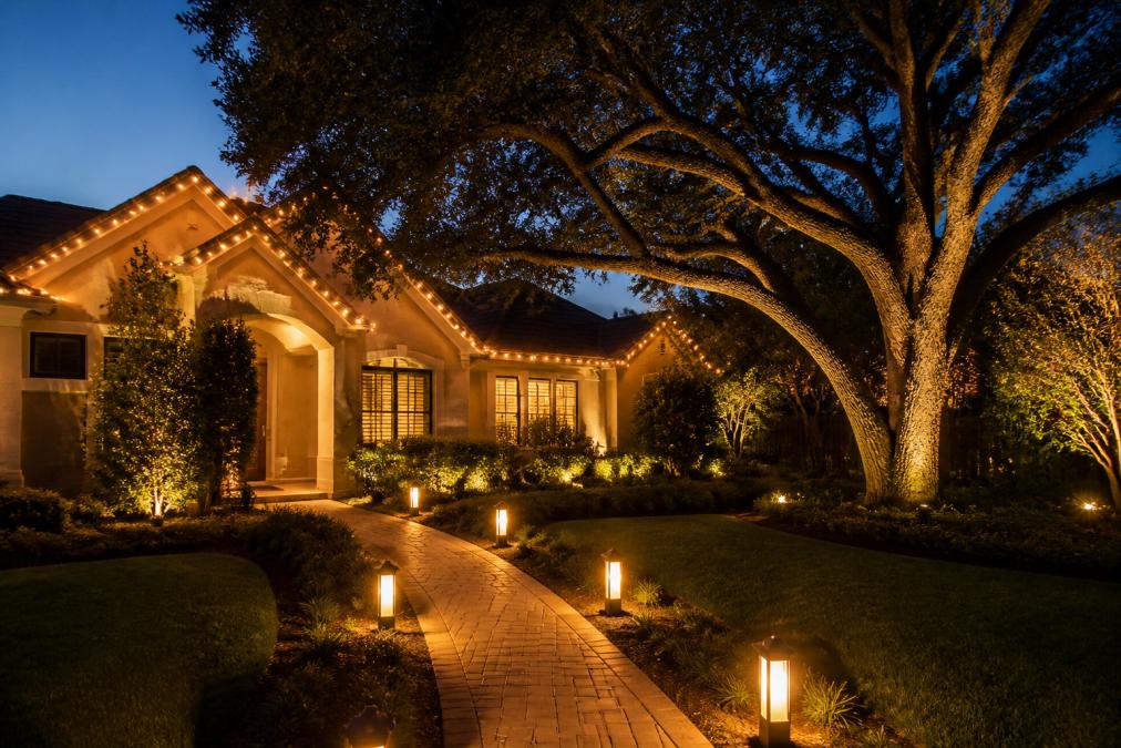 A beautifully lit residential home at dusk with warm 2700K string lights draped across the roofline, glowing pathway lanterns lining the front walkway, and landscape spotlights illuminating mature oak trees and sculpted shrubs against a deep blue evening sky
