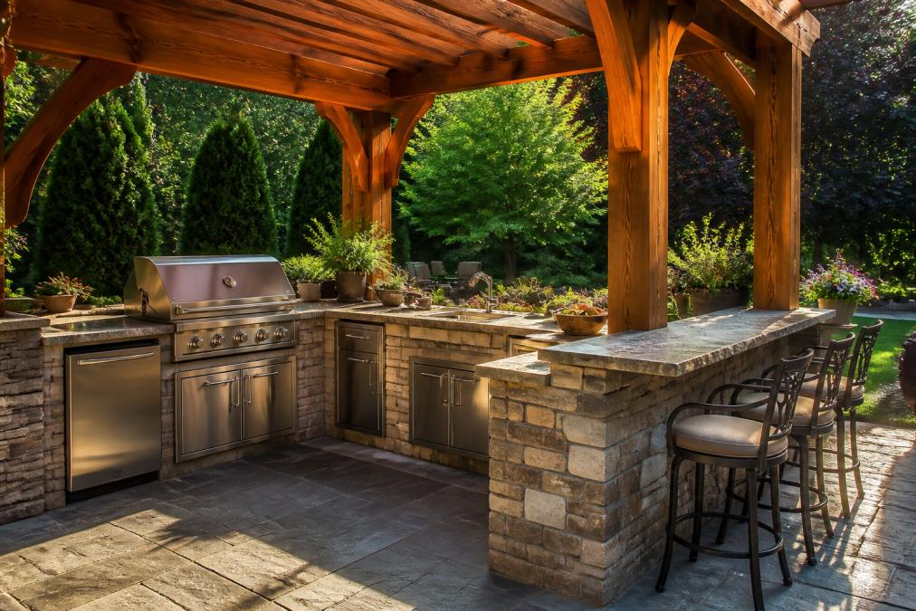 A well-organized backyard outdoor kitchen with a built-in grill station, food preparation zone, and bar seating on a stone patio, surrounded by green landscaping under a timber pergola in warm afternoon sunlight