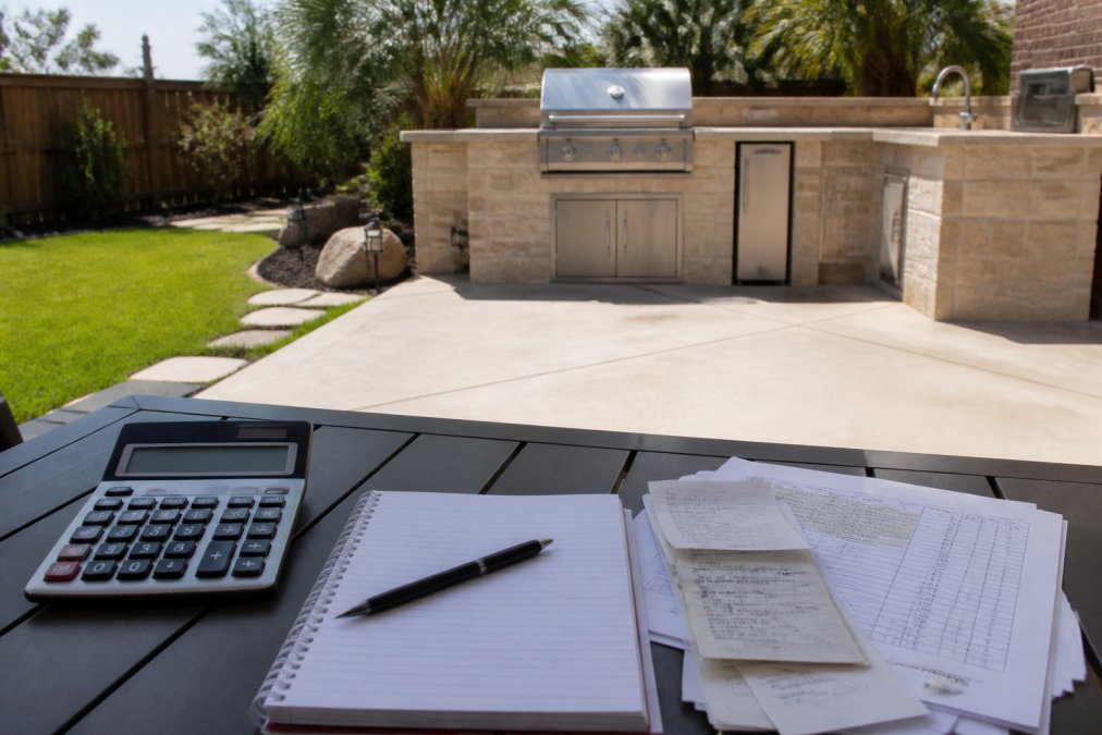 Completed outdoor kitchen patio with finished concrete slab foundation, visible utility connection points, built-in grill and stone countertops, landscaping and hardscaping elements surrounding the installation space