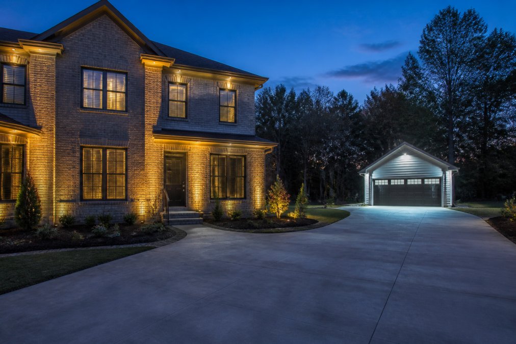 A modern two-story home at dusk with warm white 2700K lighting washing over its brick facade, while a separate detached garage in the background uses a cool blue-white 5000K floodlight, clearly illustrating the visual temperature contrast