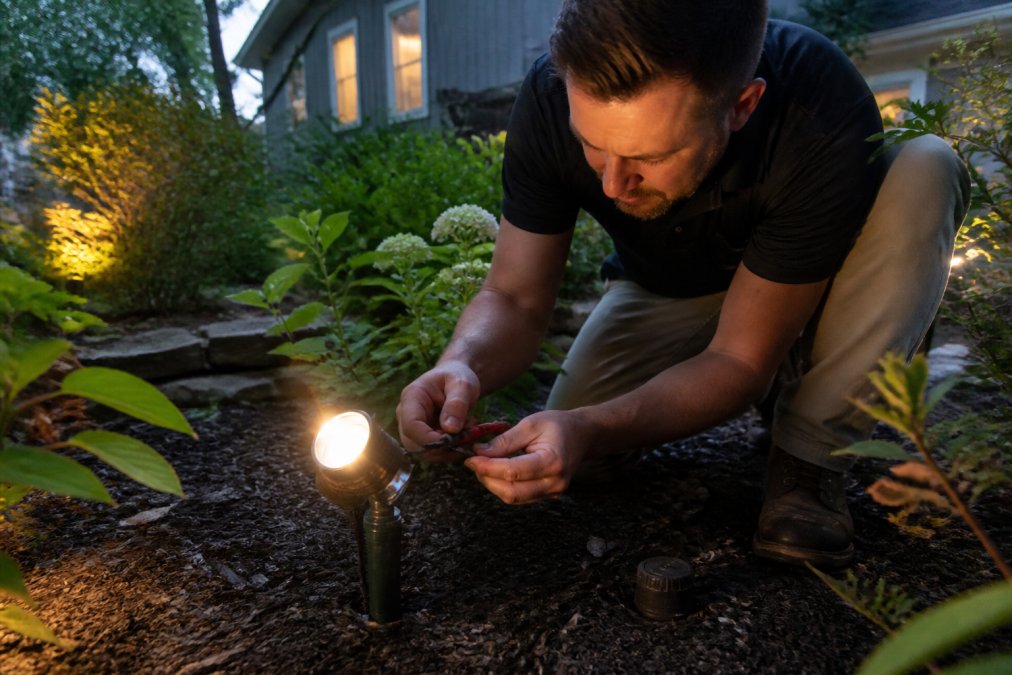 Homeowner inspecting outdoor landscape lighting fixtures at dusk, closely examining wiring connections and fixture housing for debris, corrosion, and weatherproofing integrity