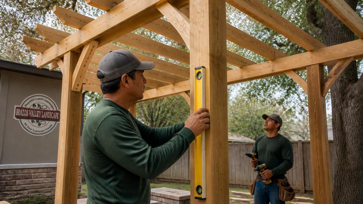 A carpenter in work clothes checking a post with a spirit level on a wooden pergola frame in a backyard, with beams and rafters visible and a second worker holding a drill nearby