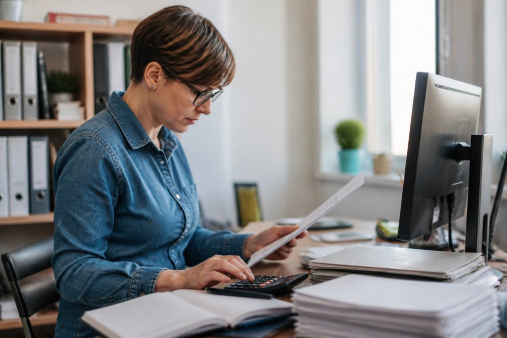 Small business owner at a desk reviewing financial reports on a computer screen with organized files, calculator, and documents nearby, showing focus and confidence in a bright office setting