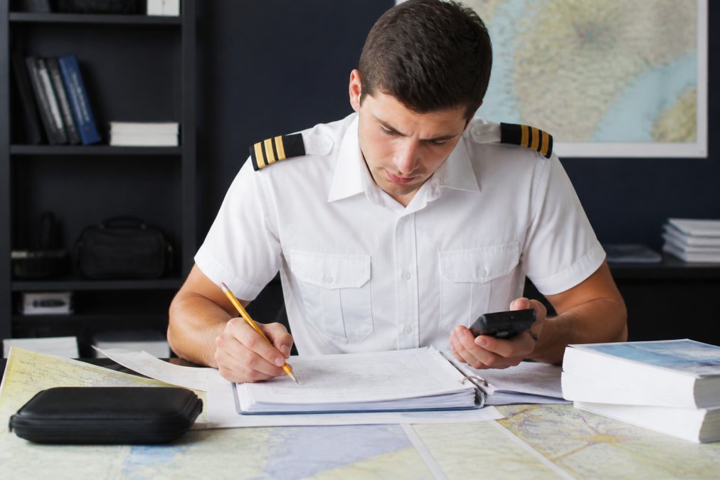 Student pilot studying flight planning calculations at a desk with aviation charts, pencil, and calculator in hand, focused expression showing concentration in natural office lighting
