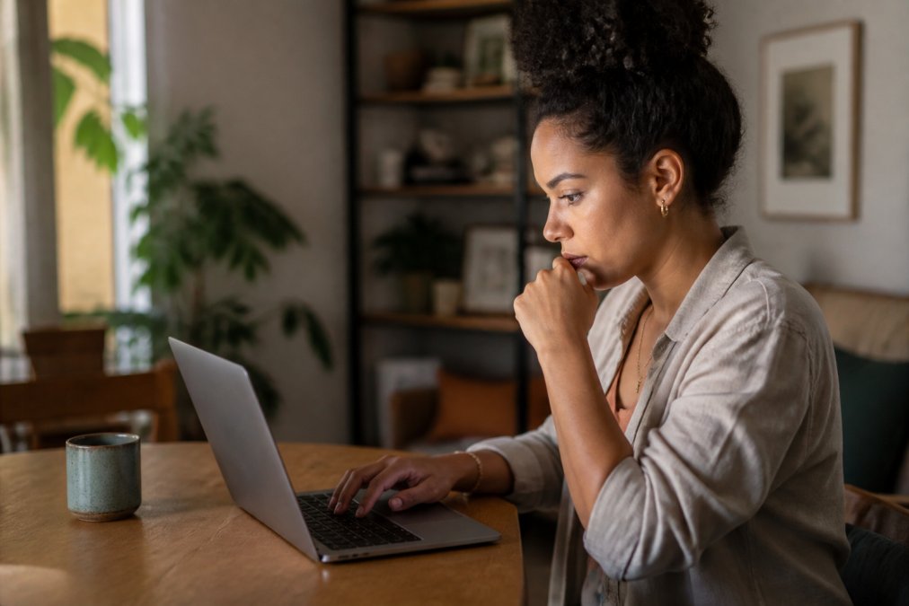 Person sitting at a laptop in a warm, comfortable home setting, looking thoughtful while typing a message in an online chat or forum interface visible on screen
