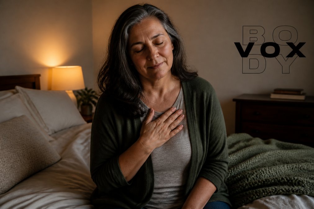 A mature person sitting alone in a softly lit bedroom, hand placed gently on their own chest, eyes closed in a moment of quiet reflection and self-awareness