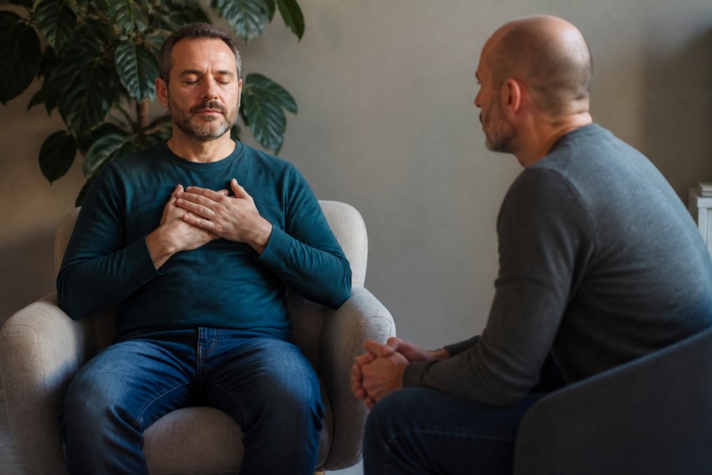 A man in his 40s sitting calmly in a softly lit therapy room, eyes closed, hands resting on his chest, with a somatic coach sitting nearby in a supportive posture