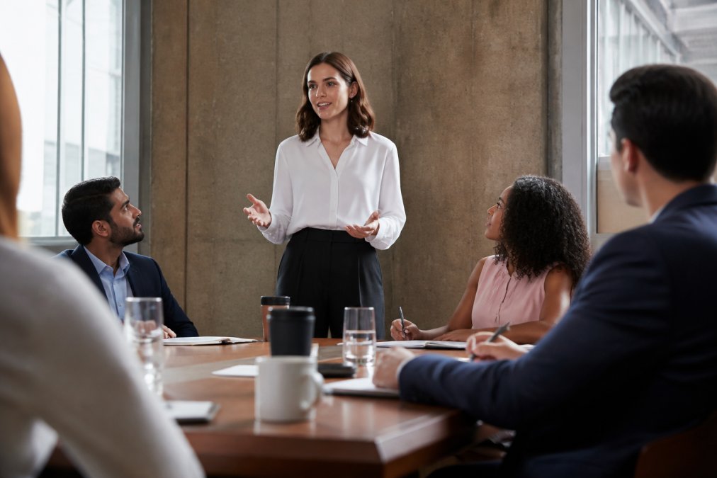 Professional speaking up confidently in a meeting room with colleagues listening attentively, demonstrating deliberate action and movement past self-doubt