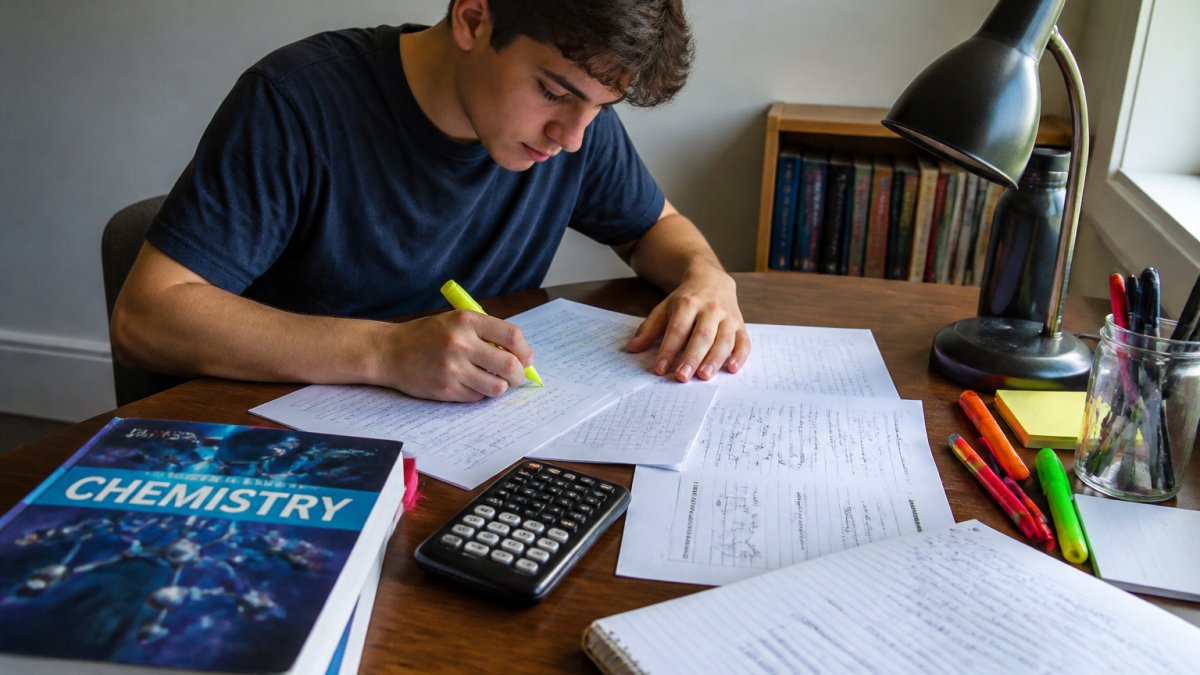 A high school student at a tidy desk covered with chemistry notes, a calculator, and a practice exam, working through problems with a highlighter in hand
