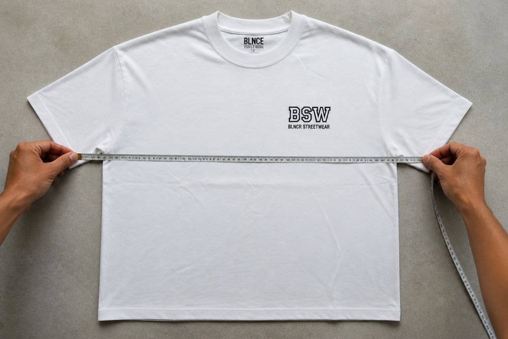 Close-up of hands using a soft measuring tape across the chest of a heavyweight cotton tee laid flat on a light grey surface, showing proper measurement technique with tape running horizontally