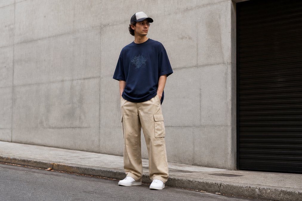 Person iført en oversized gaming-T-shirt i mørkeblå med subtil grafik, kombineret med wide-leg-cargobukser i beige, minimalistiske hvide sneakers og en truckercap, stående i en afslappet positur, der viser hele outfittet i naturligt dagslys