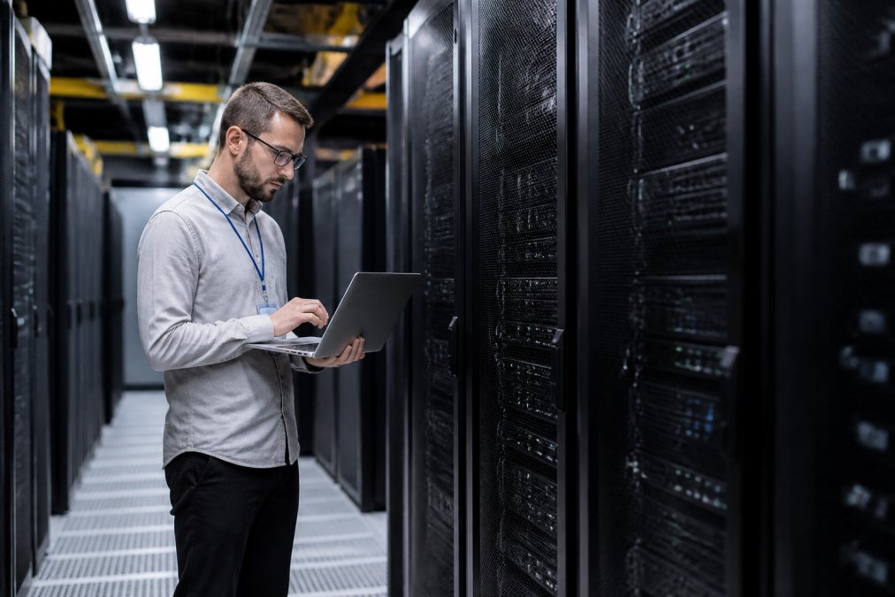 A technician in a data center aisle inspecting a rack of dedicated servers with a laptop, overhead lighting reflecting off brushed metal cabinets