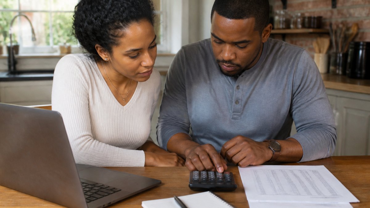 A couple sitting at a kitchen table with a calculator, laptop, and printed payment schedule, reviewing home financing numbers together in warm afternoon light