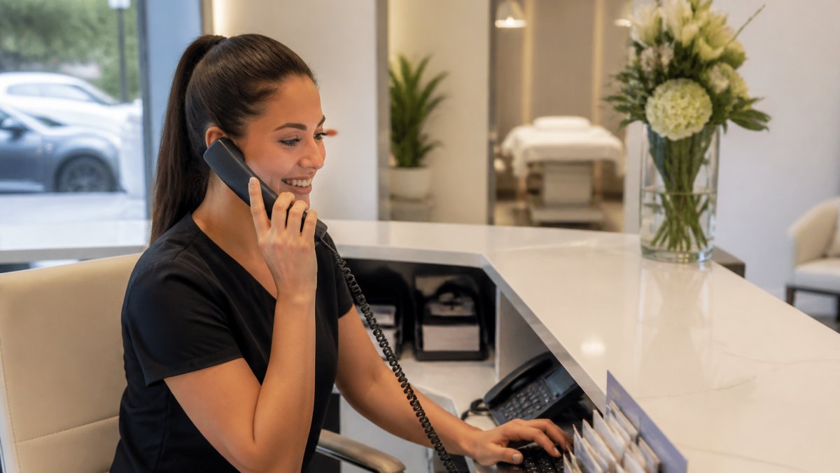 Professional med spa receptionist at desk managing patient calls and reviewing scheduling software on multiple monitors in modern clinic setting with natural light