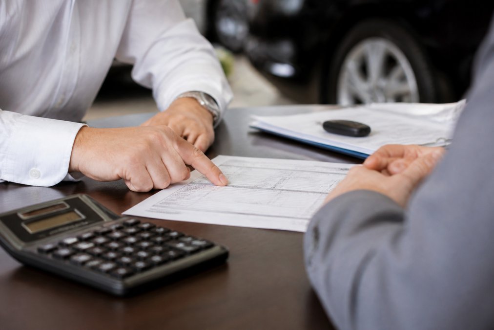 Customer reviewing detailed out-the-door pricing document at dealership desk with salesperson pointing to itemized line items and calculator visible