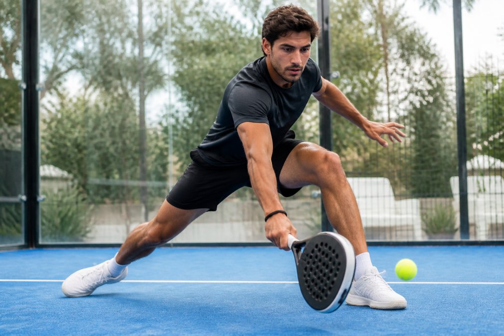 A padel player in a dynamic lunge position reaching for a low shot, wearing a fitted short-sleeve top and shorts, with a padel court and glass wall visible in the background