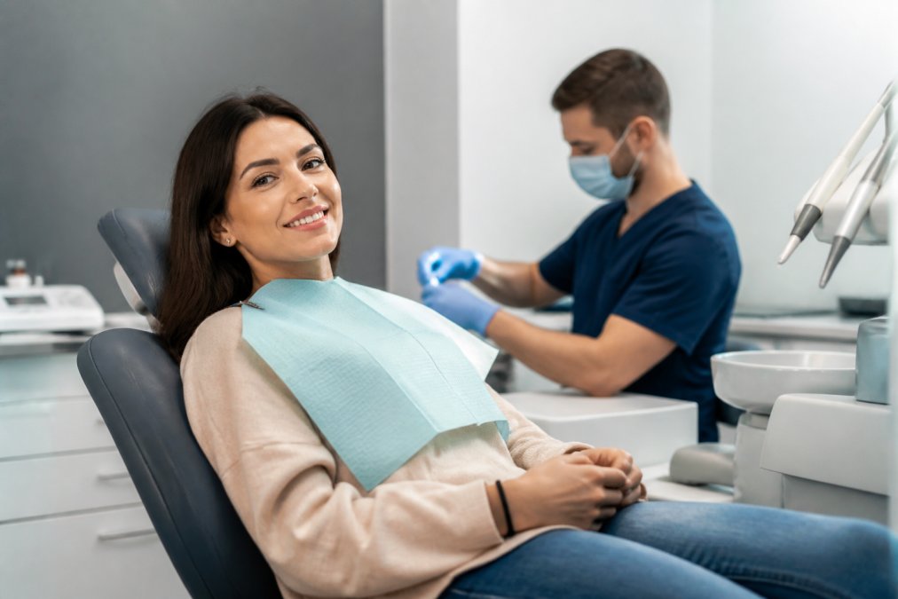 Patient sitting calmly in modern dental chair with soft clinical lighting, dentist in background preparing instruments, relaxed facial facial expression and open body language