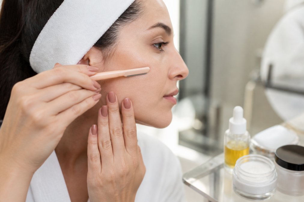 Close-up of hands performing at-home dermaplaning with proper tool angle and pressure on smooth skin, demonstrating safe technique with soothing agents nearby