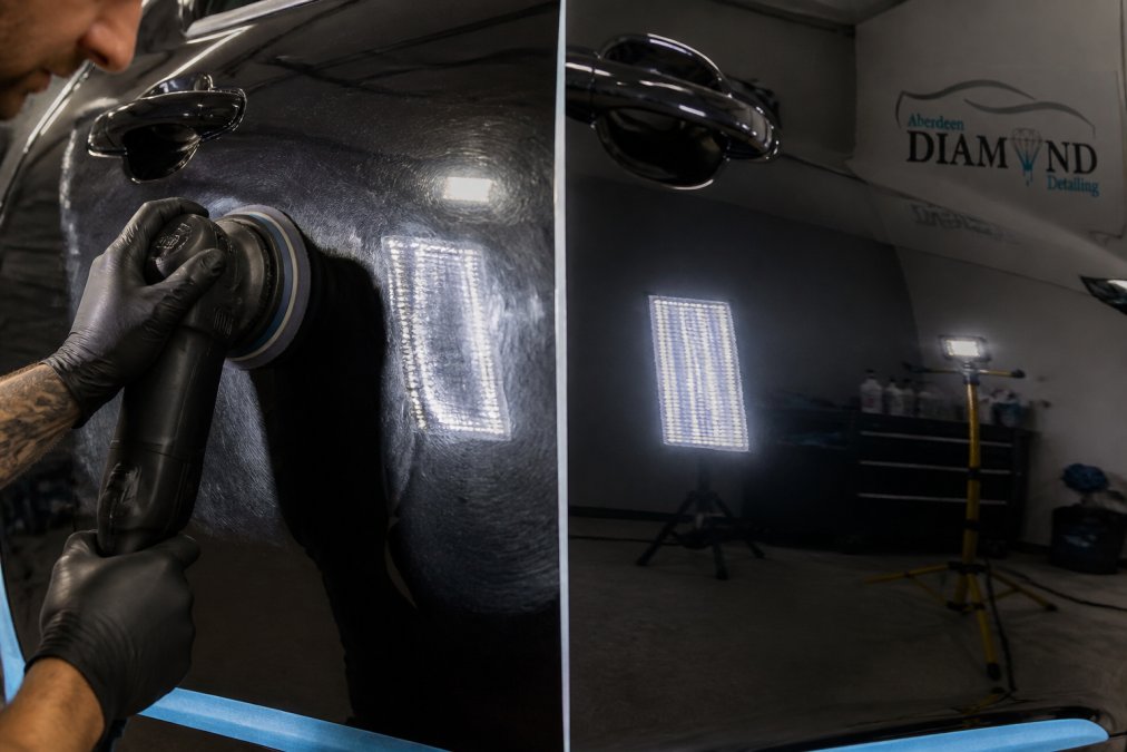 Close-up of a detailer's gloved hand holding a dual-action machine polisher against a dark car panel, with bright reflections and fine polish residue visible under LED inspection lighting in a professional detailing studio