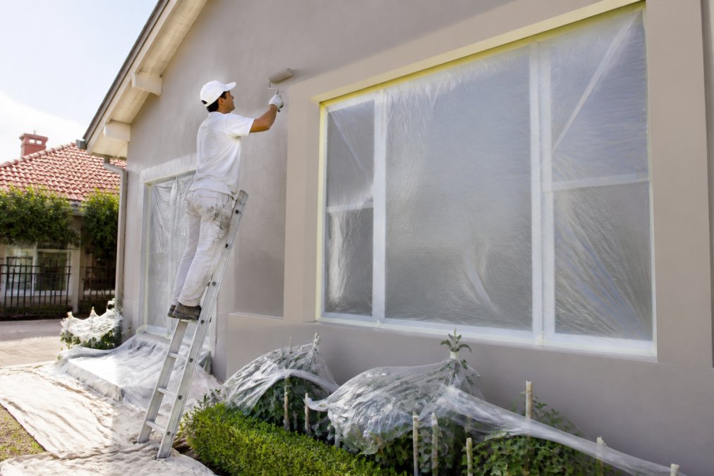 Professional painter carefully applying exterior paint to residential home facade with proper protective coverings on windows and landscaping, natural daylight illuminating the work