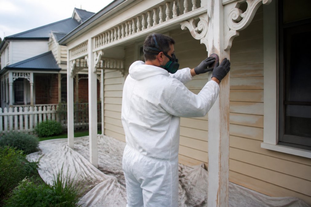 A painter in protective gear carefully sanding the decorative timber trim of a Federation-style weatherboard home, with drop sheets protecting the garden below