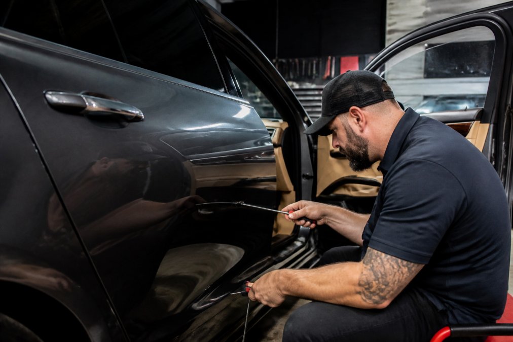 Certified PDR technician using specialized metal rods and precision tools to carefully manipulate a dent from the interior panel of a luxury vehicle, demonstrating the controlled technique in a professional repair facility