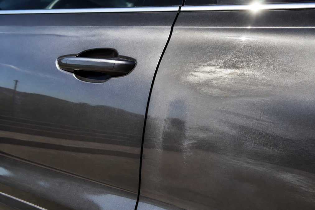Close-up of a vehicle's door panel showing original factory paint with uniform finish under natural sunlight, contrasted with a nearby section displaying visible overspray and uneven paint thickness from traditional repair work