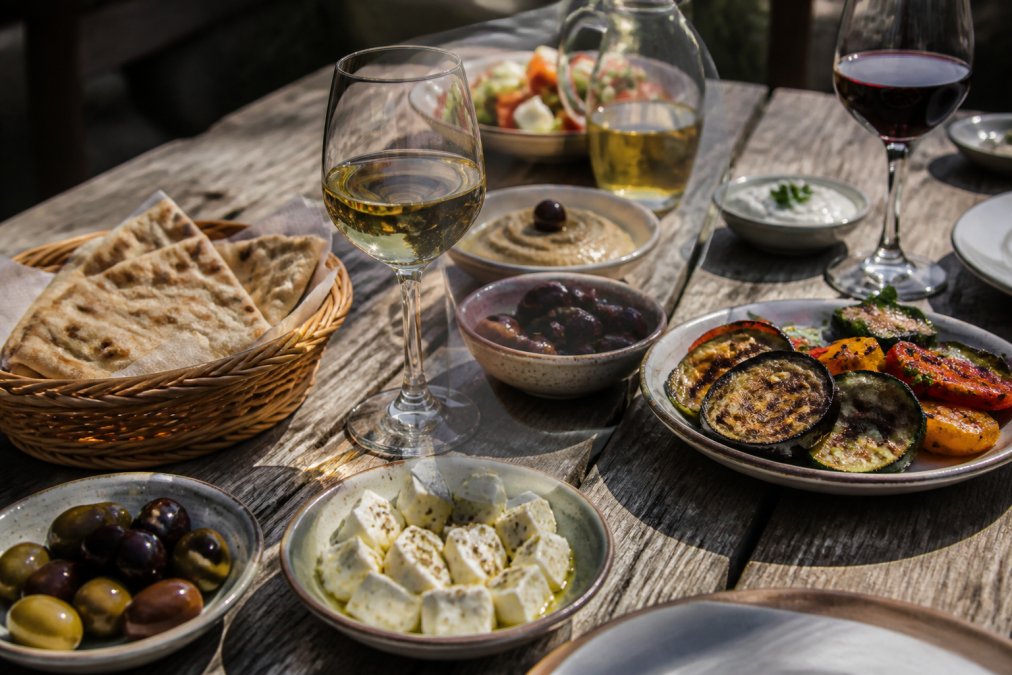 Rustic wooden table spread with Greek mezze platters including fresh pita bread, olives, feta cheese, grilled vegetables, and wine glasses filled with white and red Greek wine in warm natural light